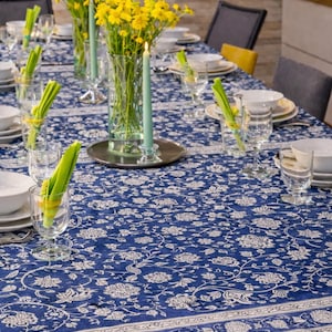 May include: A dining table set for a meal, featuring a blue floral tablecloth, white dishes, and clear glassware. Yellow flowers in vases and green candles add color. The table is set for several guests.