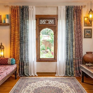 Könnte beinhalten: Ein Raum im Bohème-Stil. Ein Holzbett mit bunten Kissen und Polstern befindet sich unter einem Wandteppich. Verzierte Vorhänge umrahmen ein Fenster, und ein Teppich bedeckt den Boden. Dekorative Lampen und gerahmte Kunstwerke vervollständigen den Look.