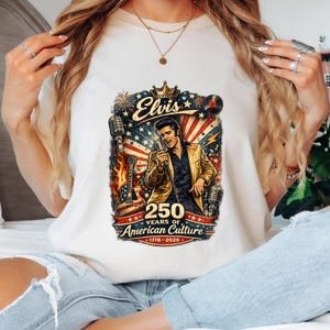 May include: Cream-colored t-shirt featuring a graphic of Elvis Presley singing into a microphone. The design includes a crown, fireworks, a guitar, and the text "Elvis" and "250 Years of American Culture 1776-2026."