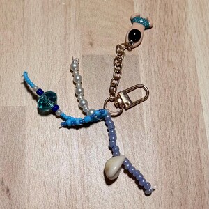 Puede incluir: Un llavero con un cierre de mosquetón y cadena dorados, unido a una pequeña figura con traje de baño azul de lunares. El accesorio incluye múltiples hilos de cuentas en tonos azules, blancos y nacarados, junto con una pequeña concha marina.