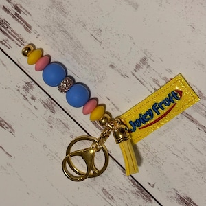 May include: A keychain featuring a row of colorful silicone beads in yellow, pink, and blue, with a central rhinestone-encrusted bead. It includes a gold-toned metal clasp, a yellow tassel, and a yellow fabric tag embroidered with the text Juicy Fruit.