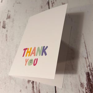 May include: A white folded greeting card featuring the text THANK YOU printed in a colorful, rounded font. The letter O in the word YOU is replaced by a light green circular smiley face icon with a simple black line for a mouth.