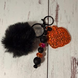 May include: A keychain featuring a black faux fur pom-pom, a black metal clasp, and a string of beads in matte black, translucent pink, and orange. An orange glittery pumpkin-shaped charm is attached, with the text Hello Pumpkin stitched on it.