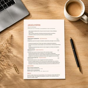 Può includere: Un curriculum vitae intitolato "Ariana Foster" con esperienza e competenze professionali, insieme a un laptop, penna, tazza di caffè e graffetta su una scrivania di legno. Il curriculum vitae descrive la storia lavorativa e l'istruzione.