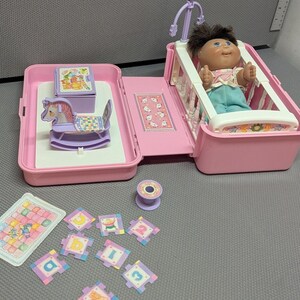 May include: A pink plastic folding doll nursery playset featuring a crib with a dark-haired doll, a rocking horse, a small storage chest, and various small accessories including letter and number tiles and a spool, all in pastel colors.