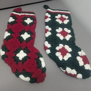 May include: Two handmade crochet Christmas stockings featuring a granny square pattern. One stocking is primarily deep red with dark green and white accents, while the other is primarily dark green with deep red and white accents. Both have a loop.