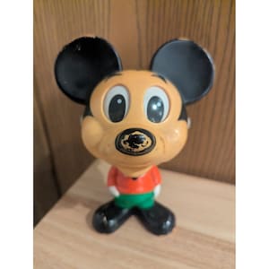 May include: A vintage Mickey Mouse figurine featuring a peach-colored face, large black ears, and white eyes with black pupils. The character wears a red shirt, green shorts, and black shoes. The black paint on the nose shows visible wear and chipping.