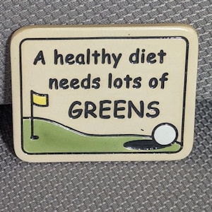 May include: A rectangular enamel pin with a cream-colored background featuring a golf course graphic with a yellow flag, a white golf ball, and a hole. The text reads: A healthy diet needs lots of GREENS in black, bold, sans-serif lettering.