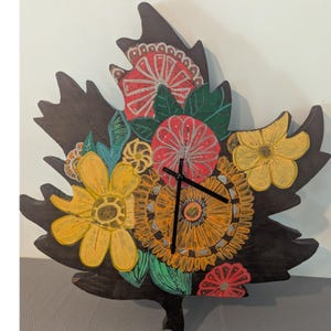 May include: A dark brown wooden wall clock shaped like a large maple leaf. The surface is decorated with hand-painted floral patterns in vibrant shades of yellow, red, orange, and green. Two black clock hands are centered on the painted flowers.