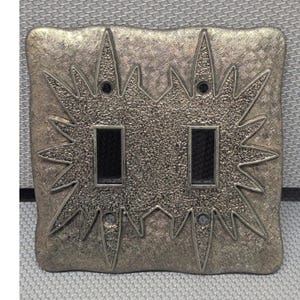 May include: A double light switch wall plate with a metallic gold finish and a textured, starburst-shaped center design. The rectangular plate features two vertical cutouts for switches and four small circular screw holes at the corners.