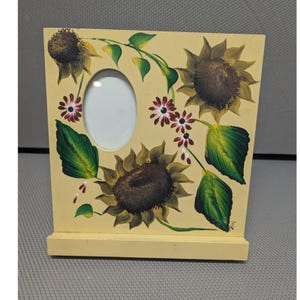 May include: A rectangular light yellow wooden picture frame featuring a central oval opening. The frame is decorated with hand-painted sunflowers in shades of brown and yellow, green leaves, and small dark red flowers on thin green stems.