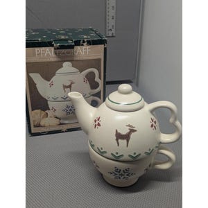 May include: A Pfaltzgraff Nordic Christmas tea-for-one set featuring a cream-colored ceramic teapot and matching cup. The pieces are decorated with brown reindeer, green chevron patterns, and blue snowflakes. The original box is included.