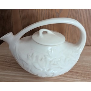 May include: A cream-colored ceramic teapot featuring a low-profile, rounded body with an embossed floral pattern. It includes a matching lid with a small handle and a single arched handle that connects the top of the spout to the back of the body.