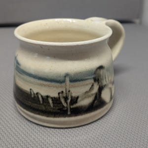 May include: A cream-colored ceramic mug with a speckled finish and a wide, rounded body. The exterior features a hand-painted desert landscape scene in muted tones of blue, brown, and black, depicting saguaro cacti and distant hills.
