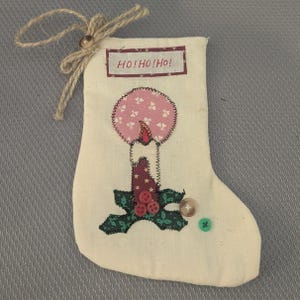 May include: A cream-colored fabric Christmas stocking featuring a stitched applique of a candle with a pink patterned flame and dark green holly leaves. The stocking includes three small red buttons, one brown button, one green button, and a jute twine tie.