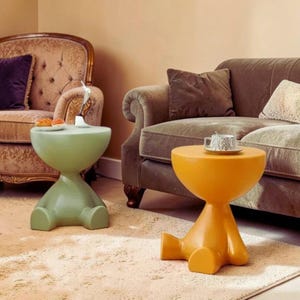 Peut inclure: Deux tables d'appoint modernes, une vert clair et une orange, chacune avec un dessus arrondi et une base en forme de jambes. La table verte a une petite sculpture et de la nourriture. La table orange a une tasse et une soucoupe.