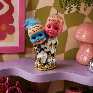 Puede incluir: Una figura caprichosa con dos cabezas de muñeca pintadas y texto. Una muñeca tiene pelo azul y una máscara, la otra una cresta. La figura está sobre una base dorada con una bola de discoteca y detalles de perlas. El fondo tiene una combinación de colores rosa y verde.