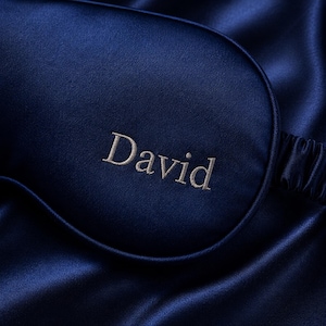 May include: A navy blue satin sleep mask embroidered with the name "David" in a light beige thread. The mask is set against a backdrop of matching satin fabric, creating a luxurious and elegant aesthetic. The sleep mask is designed to block out light.
