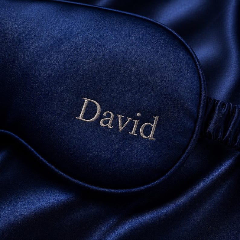 May include: A navy blue satin sleep mask embroidered with the name "David" in a light beige thread. The mask is set against a backdrop of matching satin fabric, creating a luxurious and elegant aesthetic. The sleep mask is designed to block out light.