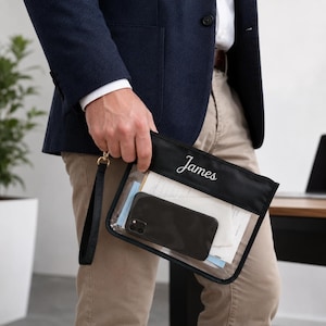 Op de afbeelding: Een zwarte en doorzichtige documententas met de naam "James" in het wit geborduurd. De tas bevat een smartphone en papieren. Hij heeft een zwarte polsband en goudkleurige hardware.