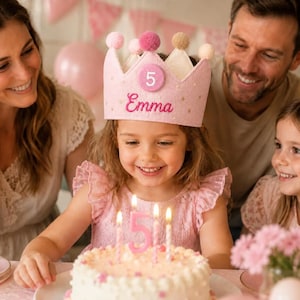 May include: A light pink felt birthday crown featuring a circular patch with the number 5, the name Emma embroidered in cursive, and small gold star accents. The crown is topped with five small pom-poms in shades of pink and beige.