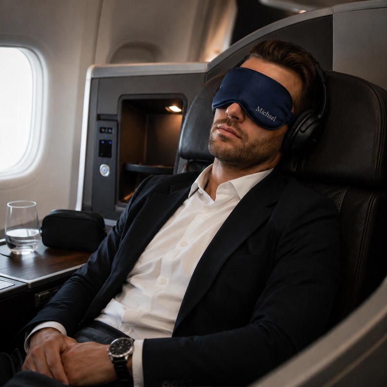May include: A navy blue sleep mask with the name "Michael" embroidered on it. The mask is worn by a person wearing a black suit and white shirt, seated in an airplane seat. The person is also wearing black headphones.