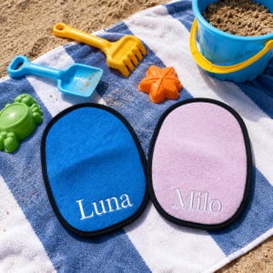 Peut inclure: Deux gants en tissu de forme ovale avec bordure noire, l'un bleu vif avec le nom Luna brodé en blanc, et l'autre violet clair avec le nom Milo brodé en blanc, posés sur une serviette de plage à rayures bleues et blanches.