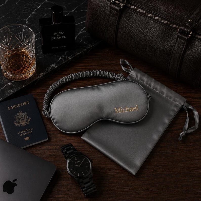 May include: A flat lay arrangement with a grey sleep mask embroidered with "Michael", a matching pouch, a black watch, a passport, a laptop, a glass of amber liquid, and a cologne bottle. A dark brown leather duffel bag is in the background.