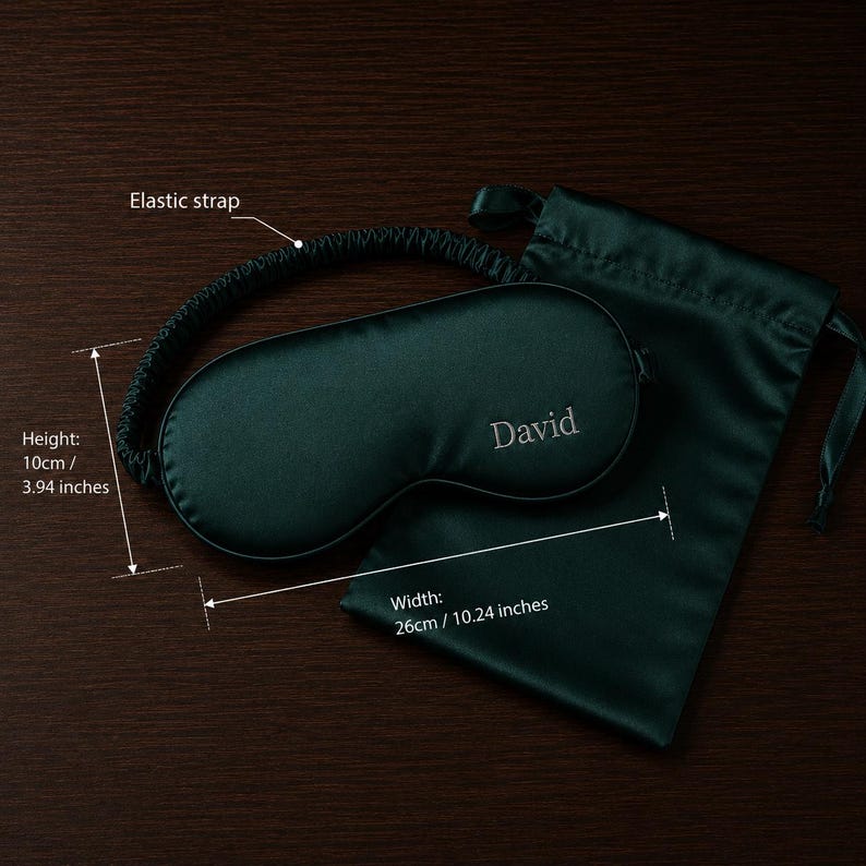 May include: A dark green satin sleep mask, embroidered with the name "David", and a matching drawstring pouch. The mask is 10cm high and 26cm wide, with an elastic strap. The set is presented on a dark brown surface.
