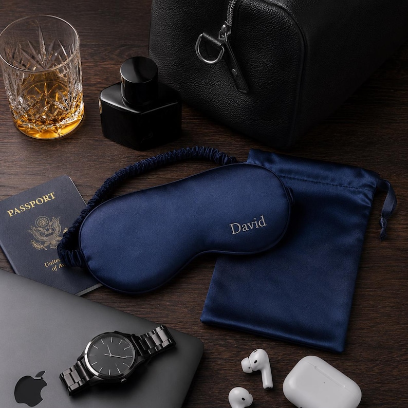 May include: A navy blue sleep mask with the name "David" embroidered on it, alongside a matching satin pouch. The image also includes a watch, passport, and a glass of amber liquid.