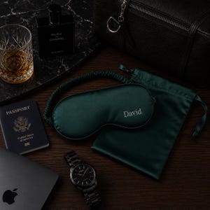 May include: A collection of travel essentials featuring a dark green sleep mask embroidered with "David", a passport, a black watch, a bottle of cologne, a glass of amber liquid, a laptop, and a black leather bag.