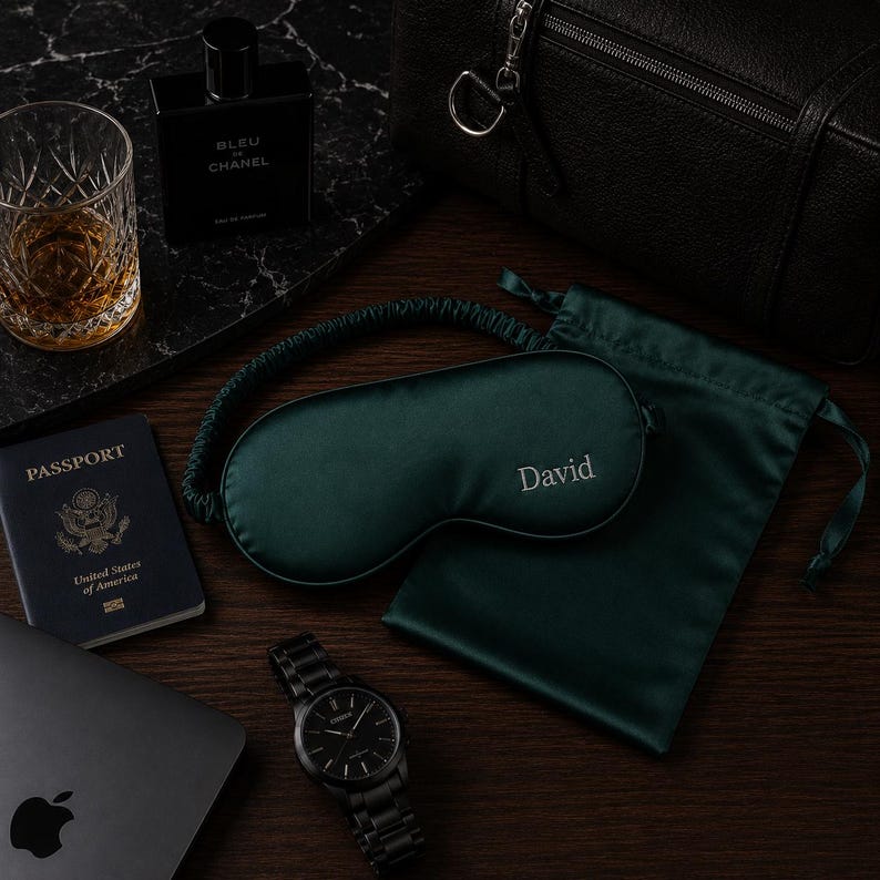 May include: A collection of travel essentials featuring a dark green sleep mask embroidered with "David", a passport, a black watch, a bottle of cologne, a glass of amber liquid, a laptop, and a black leather bag.