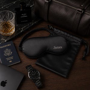 May include: A black sleep mask embroidered with the name "James", a black watch, a passport, a glass of amber liquid, and a bottle of cologne are arranged on a wooden surface. A black leather duffel bag and a black satin pouch are also present.