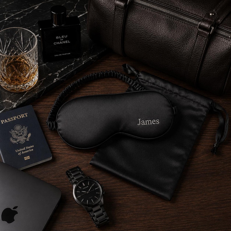 May include: A black sleep mask embroidered with the name "James", a black watch, a passport, a glass of amber liquid, and a bottle of cologne are arranged on a wooden surface. A black leather duffel bag and a black satin pouch are also present.