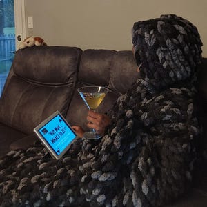 Puede incluir: Una manta con capucha de punto grueso en tonos variados de negro, carbón y gris. La manta está colocada sobre una figura sentada que sostiene una copa de martini y mira una pantalla de tableta con el texto, But Wait... What's In It?