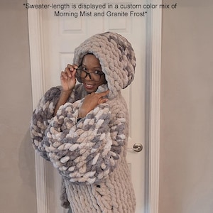 May include: A chunky knit hooded sweater in a custom color mix of Morning Mist and Granite Frost. The text on the image reads: *Sweater-length is displayed in a custom color mix of Morning Mist and Granite Frost*. The sweater has long sleeves.