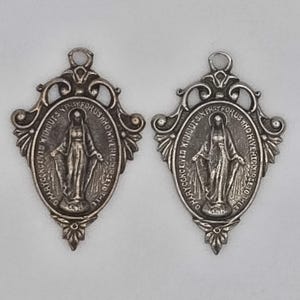 Op de afbeelding: Twee zilverkleurige ovale hangers met een religieuze figuur met uitgestrekte armen. De hangers hebben sierlijke frames met tekst rondom de centrale afbeelding. De tekst luidt: "O Maria, ontvangen zonder zonde, bid voor ons die onze toevlucht tot u nemen."