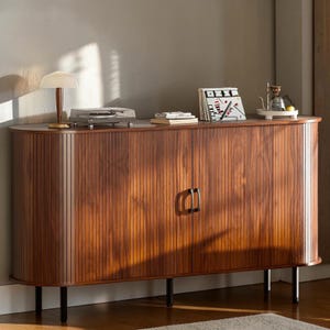 May include: A long, oval-shaped wooden sideboard featuring vertical fluted paneling, two central doors with black handles, and four slender black legs. The top surface holds a small lamp, a record player, stacked books, and a coffee dripper.