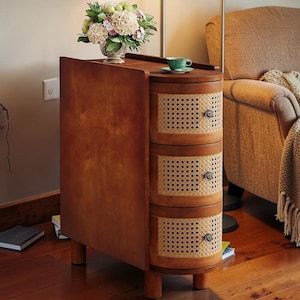 May include: A wooden side table with three drawers featuring woven cane fronts. The table has a curved design and is topped with a small green teacup. A vase of flowers sits on top. The table is 30 inches tall.