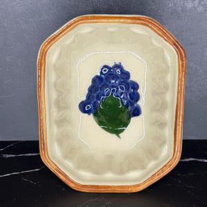 May include: A rectangular ceramic mold with a cream-colored interior and a brown-glazed rim. The center features a raised, hand-painted design of a cluster of blue grapes with a single green leaf, set against a scalloped, textured background.