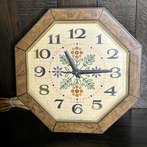 May include: Vintage General Electric wall clock with an octagonal brown frame. The clock face is cream-colored with black numerals, black hands, and floral designs in orange, green, and purple. The text "GENERAL ELECTRIC" and "MADE IN USA" are visible.