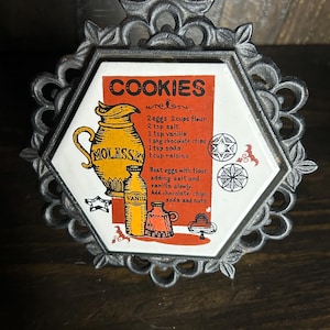 May include: A vintage metal trivet with a hexagonal ceramic tile featuring a cookie recipe. The tile has a red background with the word "COOKIES" and illustrations of a molasses jug and vanilla bottles. The metal frame has a decorative, ornate design.