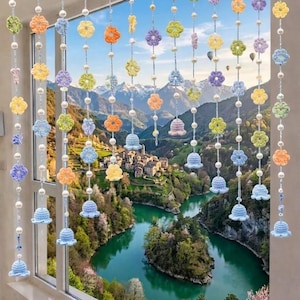 Peut inclure: Une décoration de fenêtre composée de multiples brins verticaux de fleurs crochetées et d'ornements en forme de cloche aux couleurs pastel, entremêlés de perles sphériques blanches, suspendus à une bordure en tissu bleu clair.
