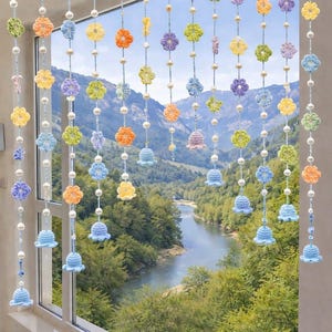 Peut inclure: Une suspension décorative pour fenêtre composée de multiples brins verticaux en fil bleu clair. Chaque brin est orné de petites fleurs crochetées multicolores dans des tons jaune, orange, vert et violet, entremêlées de perles blanches et de clochettes bleu clair.