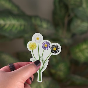 May include: A die-cut sticker featuring a bouquet of four stylized flowers with thin green stems and feathery leaves. The flowers have yellow centers and petals in white, yellow, purple, and black, arranged in a cluster on a white background.