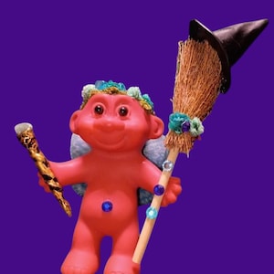 May include: A red troll figurine with a witch theme. The troll holds a wand and a broom with a black hat. The troll has blue gem accents and a floral crown. The background is a solid purple.