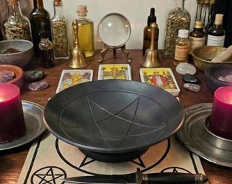 May include: A black ritual bowl with an engraved pentagram sits on a cloth featuring a pentagram design. A metal dagger with a black handle lies across the cloth. The scene includes a crystal ball, brass bells, glass bottles, tarot cards, and candles.