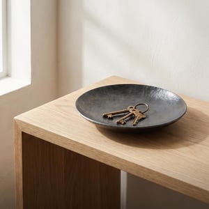 May include: A dark, circular, hand-forged metal bowl with a hammered texture. Inside the bowl rests a small metal keyring with three brass-colored keys. The text HAND-FORGED CATCHALL BOWL A RUSTIC ACCENT FOR MODERN ORGANIZATION is visible at the bottom.