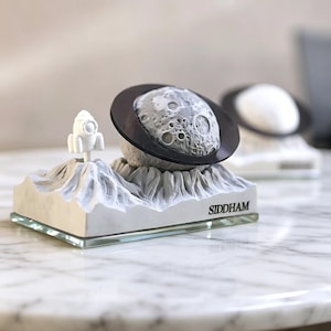 May include: A white and gray decorative sculpture featuring a moon with a black ring, a rocket, and a mountain range base. The word "SIDDHAM" is printed on the base. The sculpture is displayed on a marble table.