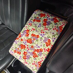 May include: A rectangular car center console armrest cover featuring a cream-colored background with a dense, repeating pattern of small red, orange, and purple flowers with green stems and leaves, fitted over a black leather vehicle console.